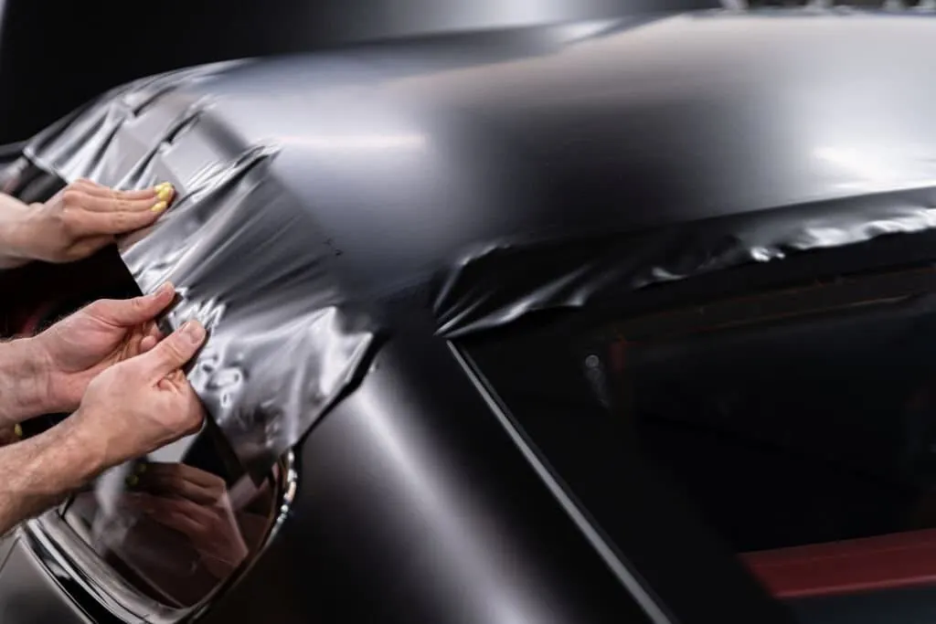 Hands smoothing satin black vinyl over a car rear quarter panel